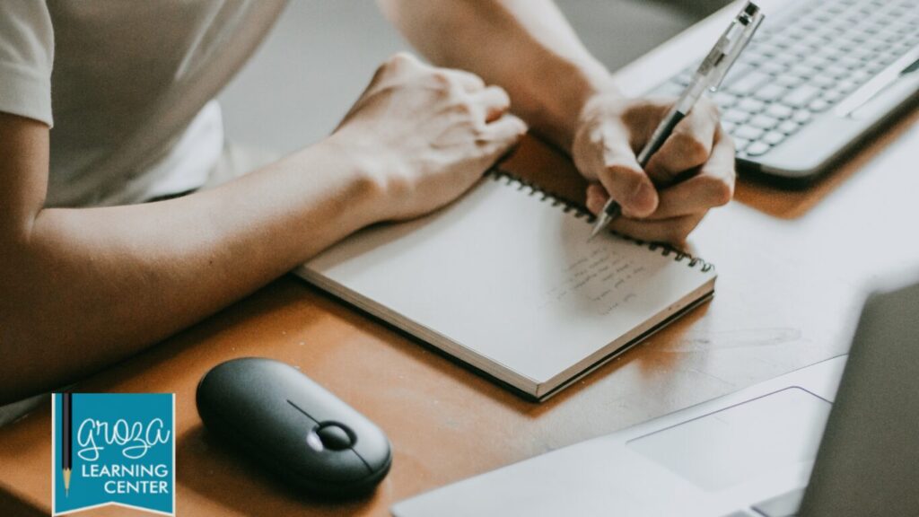 Person writing in notebook at desk with computer.